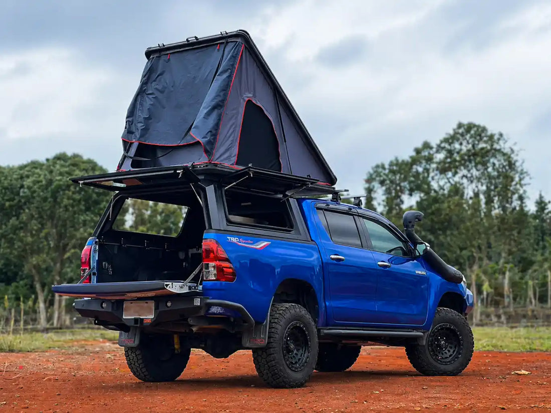 ArmaCap heavy-duty manganese steel truck canopy on a modified blue overlanding Toyota Hilux, featuring open gullwing side doors and a hardshell rooftop tent deployed, showcasing industrial-strength off-road capability.