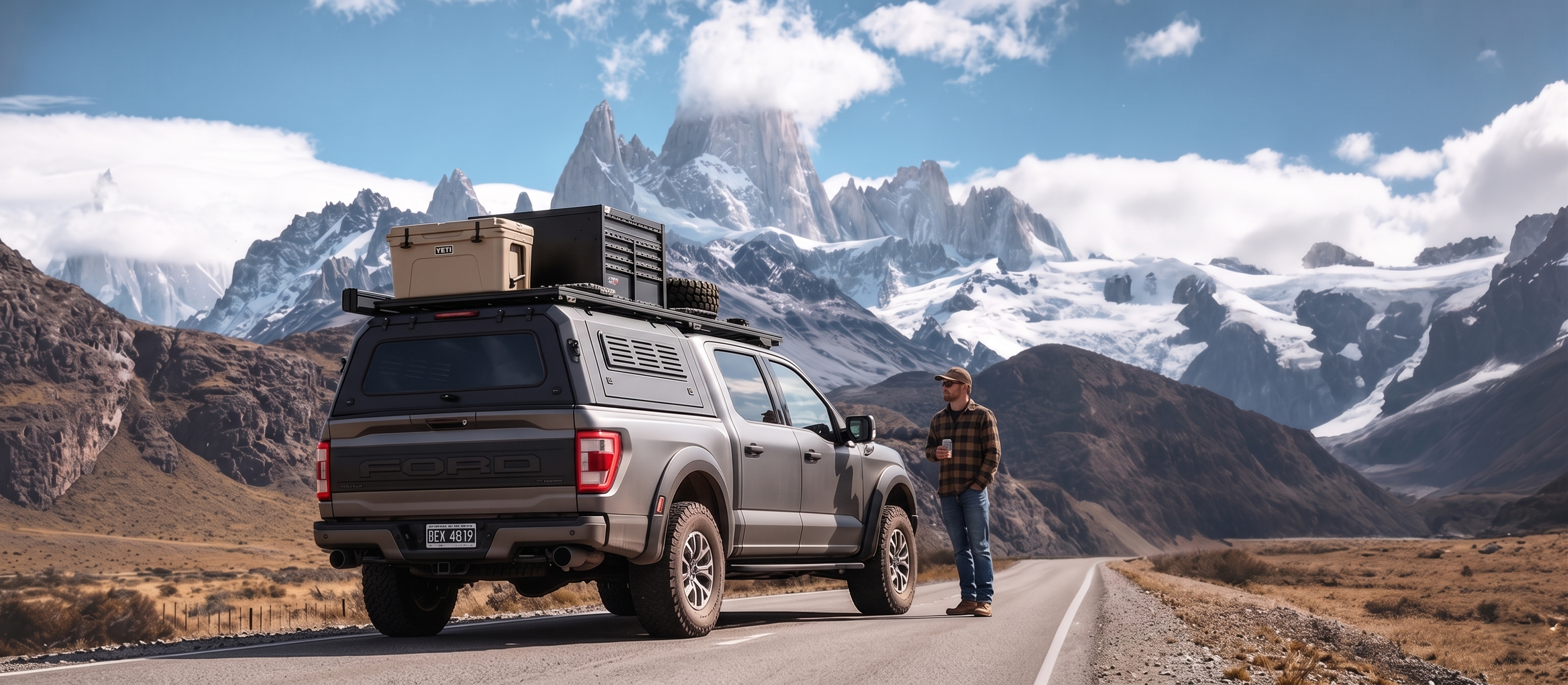 A gray Ford F‑150 pickup truck equipped with an ArmaCap heavy‑duty truck cap and roof rack is parked beside a dirt road with Mount Fitz Roy in Chile in the background. A Yeti cooler and storage box are mounted on the truck’s rear cap. A man wearing a plaid shirt stands next to the truck, gazing at the magnificent snow‑capped mountains against a bright blue sky dotted with white clouds.